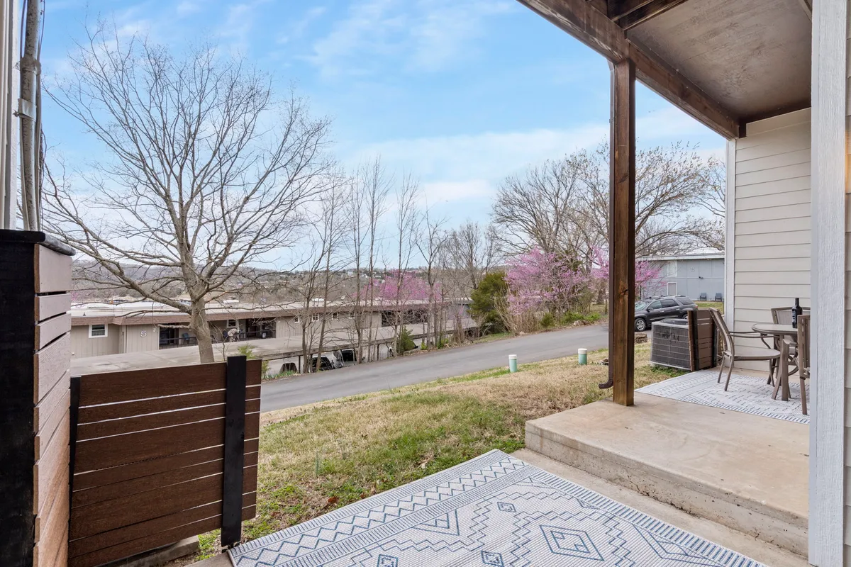 A tranquil front porch with a decorative rug, surrounded by trees and overlooking a peaceful road. The setup feels cozy and inviting, ideal for morning coffee or quiet conversation.