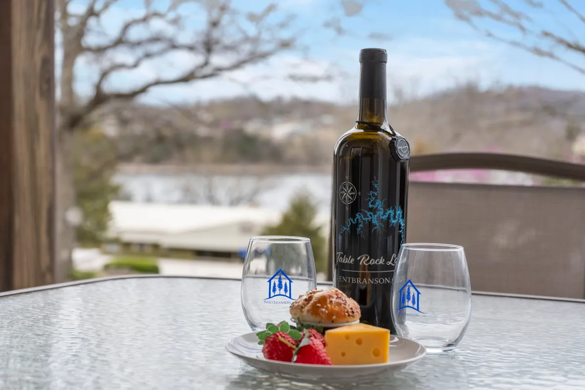 This exterior image shows a close-up of a wine bottle, two wine glasses, and a platter of food set on a table, with a scenic view of the lake in the background. The setup feels casual, indulgent, and romantic, emphasizing the enjoyment of local food and wine in a beautiful natural setting.