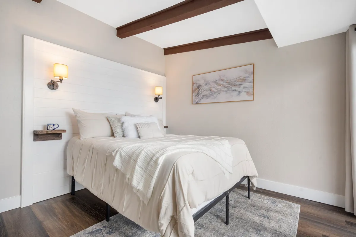 This image features a tranquil bedroom setting with a neatly made bed, wall art, and a nightstand. Soft lighting and neutral tones create a calm and inviting atmosphere, perfectly suited for relaxation and restful sleep.