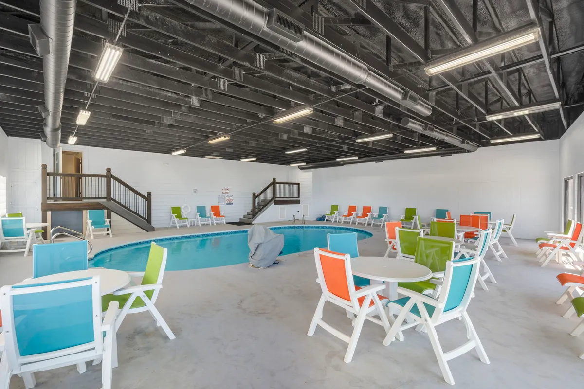 A cheerful indoor pool area with vivid chairs and tables around a sparkling swimming pool, bathed in natural light. The space is designed for year-round fun regardless of the weather.