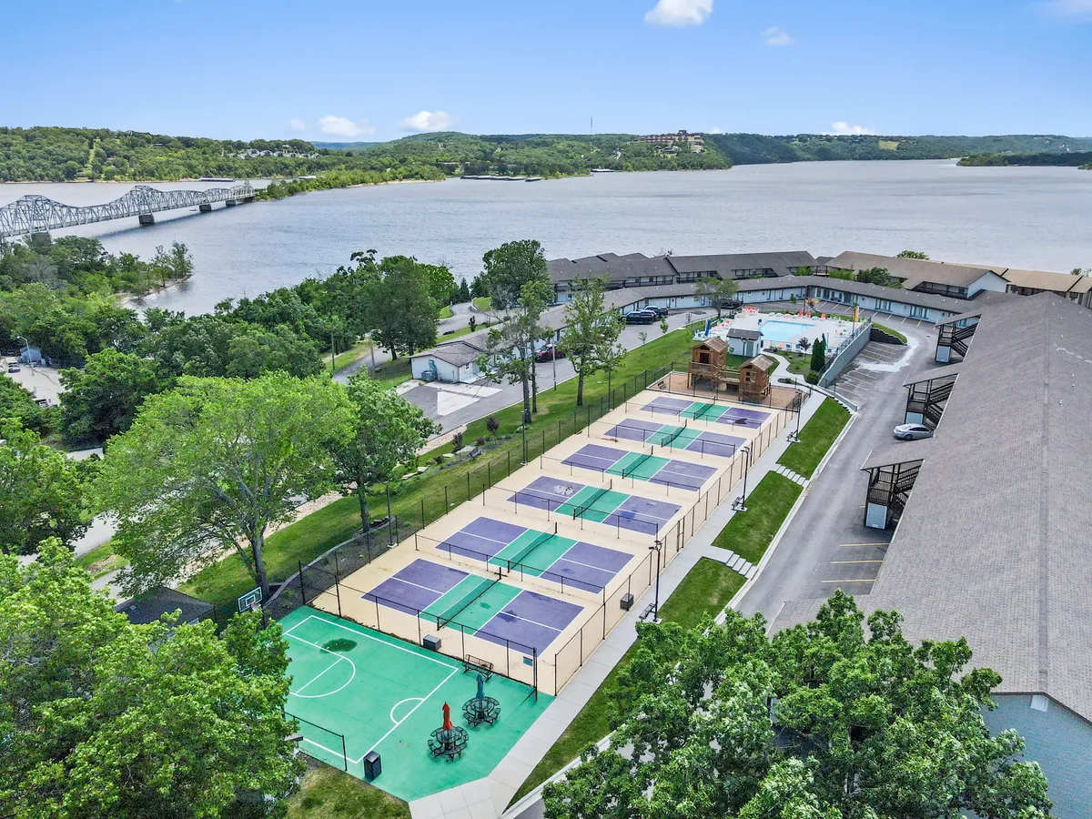 A wide exterior view of the community’s recreational area, highlighting tennis courts, a swimming pool, playground, lush greenery, and a scenic river backdrop. Families and groups can be seen enjoying various activities.