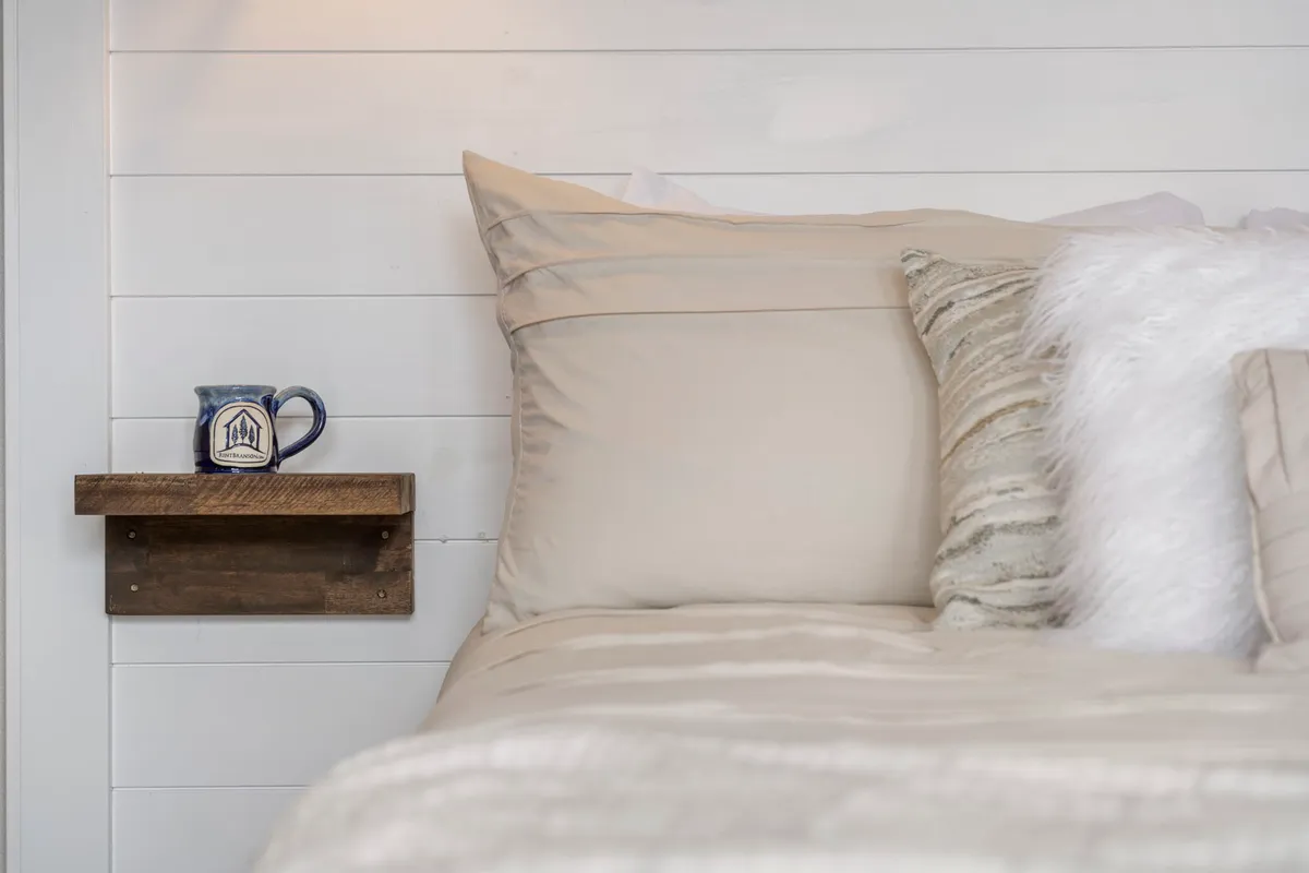 A cozy bed with soft linens, decorative pillows, and a wooden bedside shelf topped with a ceramic mug. The scene suggests a peaceful morning or evening, with an emphasis on comfort and the pleasures of solitude.
