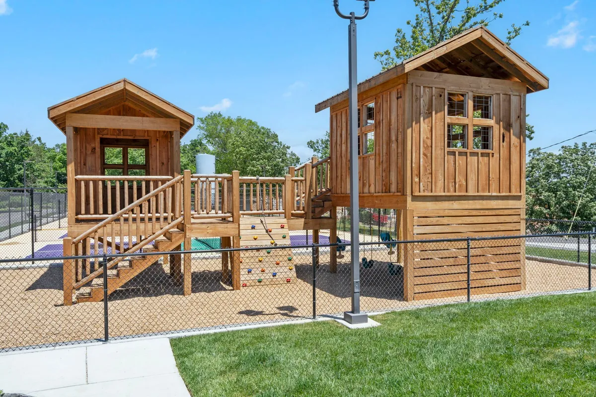 A dedicated play area with colorful playhouse structures, a climbing wall, a slide, and secure fencing, filled with children engaging in imaginative play and social interaction.